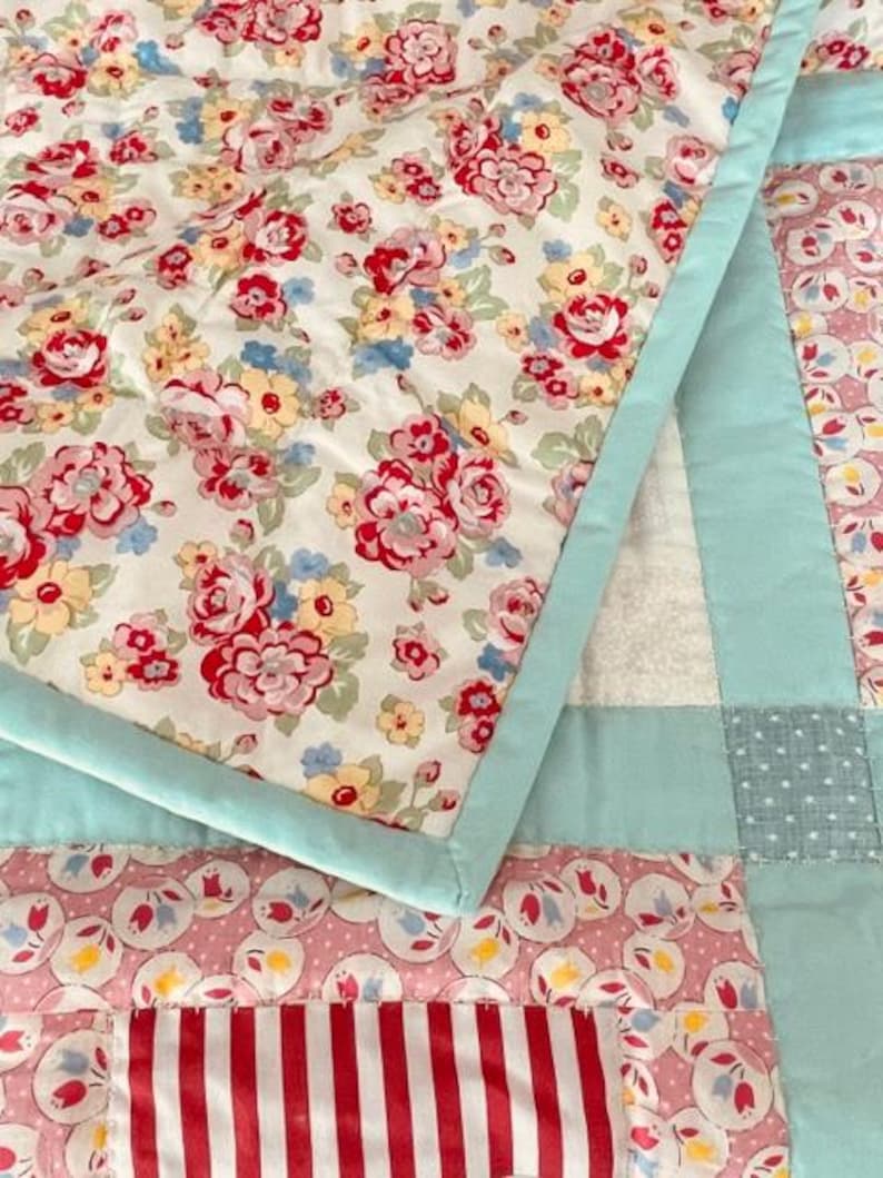 K&ouml;nnte beinhalten: Eine Patchwork-Decke mit einem Blumendruck auf der obersten Schicht. Die Decke besteht aus verschiedenen Stoffen in den Farben Rosa, Blau und Wei&szlig;. Die Decke ist mit wei&szlig;em Garn gesteppt.