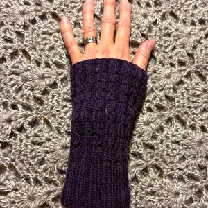 May include: A dark purple knitted wrist warmer with a textured pattern. The wrist warmer is made of a soft, warm material. The background is a gray crocheted blanket.