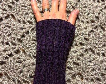 Könnte beinhalten: Ein dunkelviolett gestrickter Handgelenkwärmer mit strukturiertem Muster. Der Handgelenkwärmer besteht aus einem weichen, warmen Material. Der Hintergrund ist eine graue Häkeldecke.