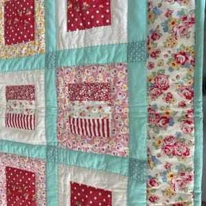K&ouml;nnte beinhalten: Eine Patchwork-Decke mit einem t&uuml;rkisfarbenen Rand und einem Blumendruck auf der rechten Seite. Die Decke besteht aus verschiedenen Stoffen mit roten, wei&szlig;en und rosa Blumenmustern.