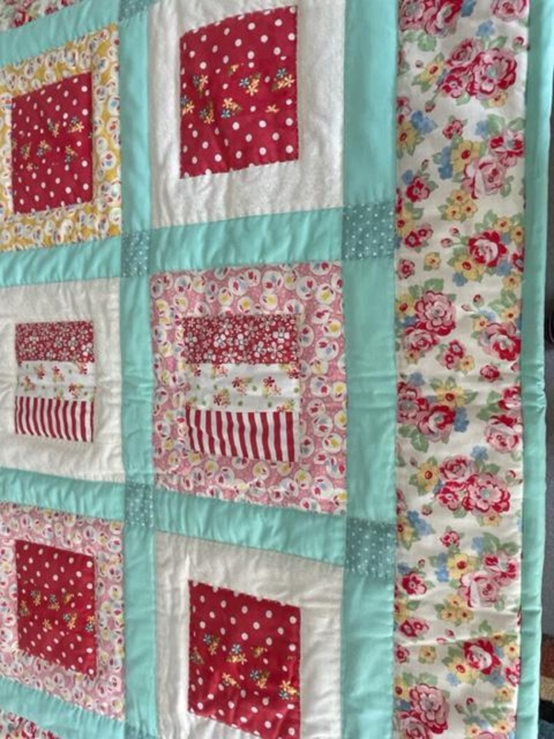 K&ouml;nnte beinhalten: Eine Patchwork-Decke mit einem t&uuml;rkisfarbenen Rand und einem Blumendruck auf der rechten Seite. Die Decke besteht aus verschiedenen Stoffen mit roten, wei&szlig;en und rosa Blumenmustern.