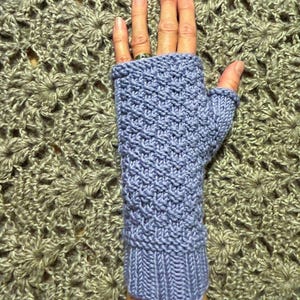 May include: A pair of purple fingerless gloves with a textured knit pattern. The gloves are shown on a hand against a crocheted background.