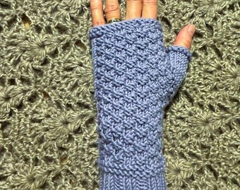 Könnte beinhalten: Ein Paar violette Fingerlose Handschuhe mit einem strukturierten Strickmuster. Die Handschuhe werden auf einer Hand vor einem gehäkelten Hintergrund gezeigt.