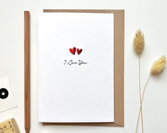 Puede incluir: Una tarjeta blanca con dos corazones rojos brillantes y las palabras "I Love You" en escritura negra. La tarjeta está dentro de un sobre marrón. Un lápiz, una goma de borrar, un clip y un sacapuntas también están en la superficie blanca.