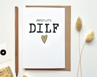 May include: A white card with black text that reads "ABSOLUTE DILF" with a gold glitter heart below the text.