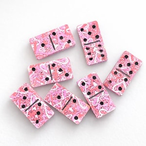 May include: A set of seven pink dominoes with a swirling pattern of white and darker pink. Each domino has black dots indicating the numbers, and a black line divides each piece. The dominoes are arranged on a white surface.