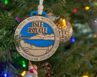Könnte beinhalten: Weihnachtsschmuck aus Holz mit blauem und beigem Design. Der Schmuck zeigt den Text "Isle Royale" und andere Wörter wie "Majestic Wilderness" und "Breathtaking". Der Schmuck hängt an einem Weihnachtsbaum.