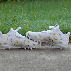 Puede incluir: Un par de tacos de f&uacute;tbol blancos con un patr&oacute;n de pl&aacute;tano. Los tacos tienen cordones amarillos y una franja amarilla en la parte posterior del tal&oacute;n.