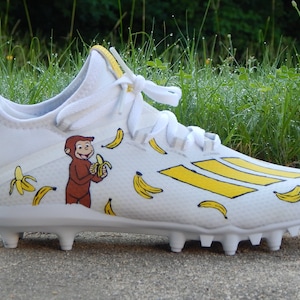 Puede incluir: Botas de f&uacute;tbol blancas con rayas amarillas y un dise&ntilde;o pintado a mano con Curious George comiendo un pl&aacute;tano. Las botas est&aacute;n cubiertas de pl&aacute;tanos amarillos.