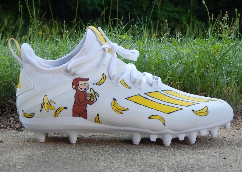 Puede incluir: Botas de f&uacute;tbol blancas con rayas amarillas y un dise&ntilde;o pintado a mano con Curious George comiendo un pl&aacute;tano. Las botas est&aacute;n cubiertas de pl&aacute;tanos amarillos.