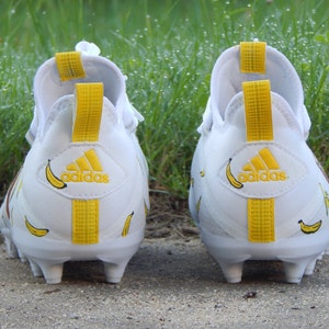 Puede incluir: Un par de tacos de f&uacute;tbol Adidas blancos con detalles amarillos y estampado de pl&aacute;tano. Los tacos tienen una franja amarilla en la parte posterior y el logotipo de Adidas en el tal&oacute;n.