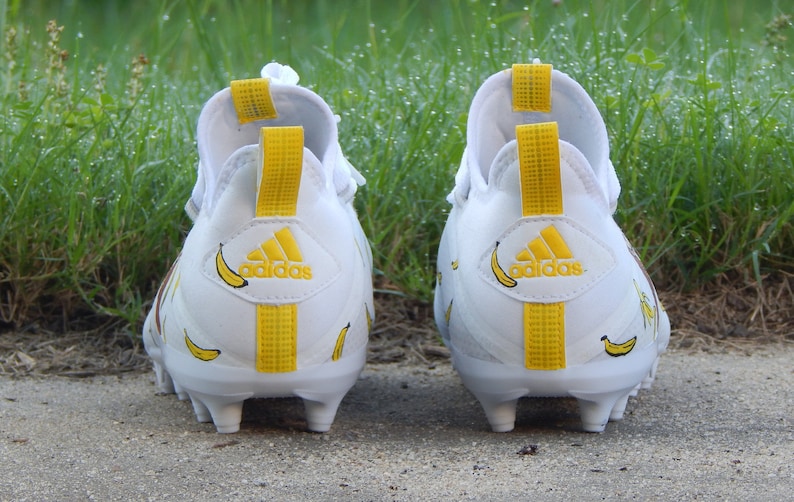 Puede incluir: Un par de tacos de f&uacute;tbol Adidas blancos con detalles amarillos y estampado de pl&aacute;tano. Los tacos tienen una franja amarilla en la parte posterior y el logotipo de Adidas en el tal&oacute;n.