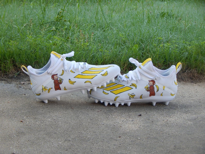 Puede incluir: Un par de tacos de f&uacute;tbol blancos con detalles amarillos y un dise&ntilde;o de mono de dibujos animados. Los tacos tienen una franja amarilla en el lateral y est&aacute;n cubiertos de pl&aacute;tanos amarillos y monos.