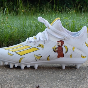 Puede incluir: Botas de f&uacute;tbol blancas con un patr&oacute;n de pl&aacute;tanos amarillos y un mono de dibujos animados comiendo un pl&aacute;tano. El mono es marr&oacute;n y tiene un pl&aacute;tano amarillo en la mano.
