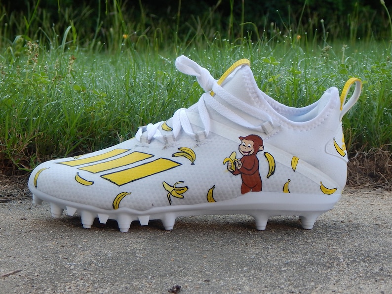 Puede incluir: Botas de f&uacute;tbol blancas con un patr&oacute;n de pl&aacute;tanos amarillos y un mono de dibujos animados comiendo un pl&aacute;tano. El mono es marr&oacute;n y tiene un pl&aacute;tano amarillo en la mano.