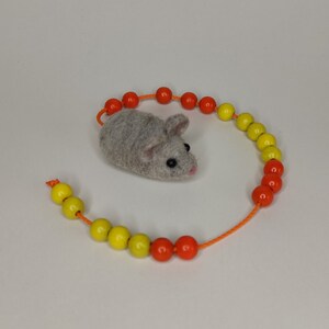 Könnte beinhalten: Ein graues Filz-Mausspielzeug mit schwarzen Augen, umgeben von einer Schnur mit orangefarbenen und gelben Perlen.