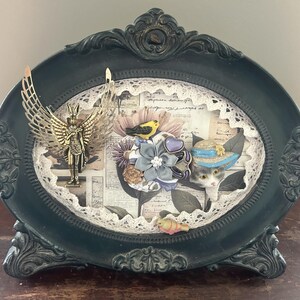 May include: An oval dark green ornate frame containing a collage with a gold-toned winged figure, a fabric flower cluster, a small yellow bird, and a cat figurine wearing a blue-trimmed hat, all set against a background of lace and script.