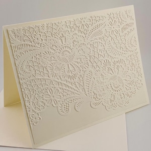 May include: Ivory-colored greeting card with a detailed, embossed lace design. The card is open, revealing the inside, and rests on a matching envelope. The intricate floral and geometric patterns create a delicate, elegant aesthetic.