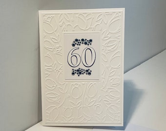 May include: A white card with a raised embossed design of leaves and flowers. The number 60 is centered on the card in a black font with a blue border.