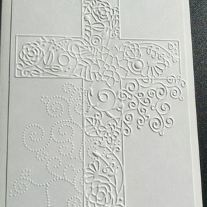 May include: A white card with an embossed cross design. The cross is decorated with intricate floral patterns and swirls.