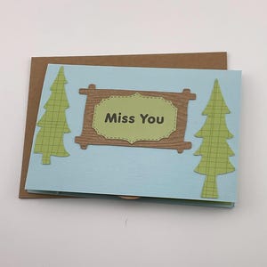 May include: A handmade greeting card with a light blue background and a brown envelope. The card features a brown wooden sign with the words "Miss You" in the center, flanked by two green tree cutouts.
