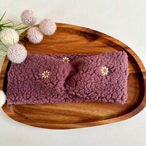 Könnte beinhalten: Ein mauvefarbenes, flauschiges Stirnband im Sherpa-Stil mit gedrehtem Vorderteil und kleinen, aufgestickten weißen Gänseblümchenmotiven mit gelben Zentren, die über die strukturierte, plüschige Stoffoberfläche verteilt sind.