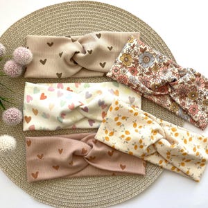 Könnte beinhalten: Fünf verschiedene Stirnbänder mit einem gedrehten Knoten-Design. Die Stirnbänder weisen verschiedene Muster auf, darunter Herzen, Blumendrucke und Zitronenmuster. Die Farben sind Beige, Rosa und Creme.