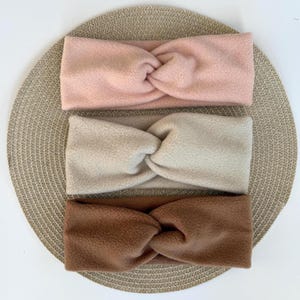 Könnte beinhalten: Drei weiche Fleece-Stirnbänder, die vertikal übereinander liegen und jeweils ein zentrales Knotendetail aufweisen. Das obere Stirnband ist hellrosa, das mittlere cremefarben und das untere Stirnband ist warmbraun, alle aus strukturiertem Stoff.