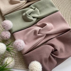 Könnte beinhalten: Gezeigt werden vier gerippte Strickstirnbänder in Beige, Grün, Rosa und Mauve. Die Stirnbänder haben ein Design mit gedrehtem Knoten. Dekorative florale Akzente sind in der Nähe der Stirnbänder platziert und verleihen der Präsentation einen Hauch von Eleganz.
