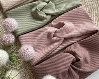 Könnte beinhalten: Gezeigt werden vier gerippte Strickstirnbänder in Beige, Grün, Rosa und Mauve. Die Stirnbänder haben ein Design mit gedrehtem Knoten. Dekorative florale Akzente sind in der Nähe der Stirnbänder platziert und verleihen der Präsentation einen Hauch von Eleganz.