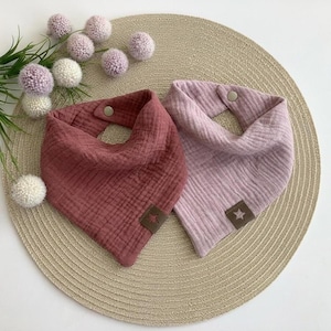 Könnte beinhalten: Zwei Spucktücher aus Musselin in Rosa und Lila, mit einem sternförmigen Aufnäher. Die Tücher sind dreieckig und haben einen Druckknopfverschluss am Hals. Auf einer beigen Matte mit Pompons.