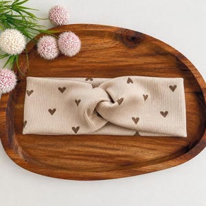 Könnte beinhalten: Ein beiges, geripptes Stoffstirnband mit einem sich wiederholenden Muster aus kleinen braunen Herzen. Das Stirnband hat ein gedrehtes Knotendetail in der Mitte und wird horizontal präsentiert, wobei die weiche Textur und das Herzmuster sichtbar sind.