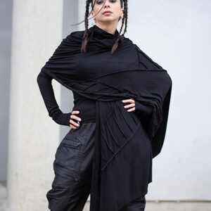 K&ouml;nnte beinhalten: Eine Frau in einem schwarzen Ensemble. Das Outfit umfasst ein lang&auml;rmliges Oberteil, einen drapierten Schal und eine dunkle Cargo-Hose aus Denim. Der Schal hat ein einzigartiges Nahtdesign. Die Frau hat dunkles Haar, das in Z&ouml;pfen gestylt ist.