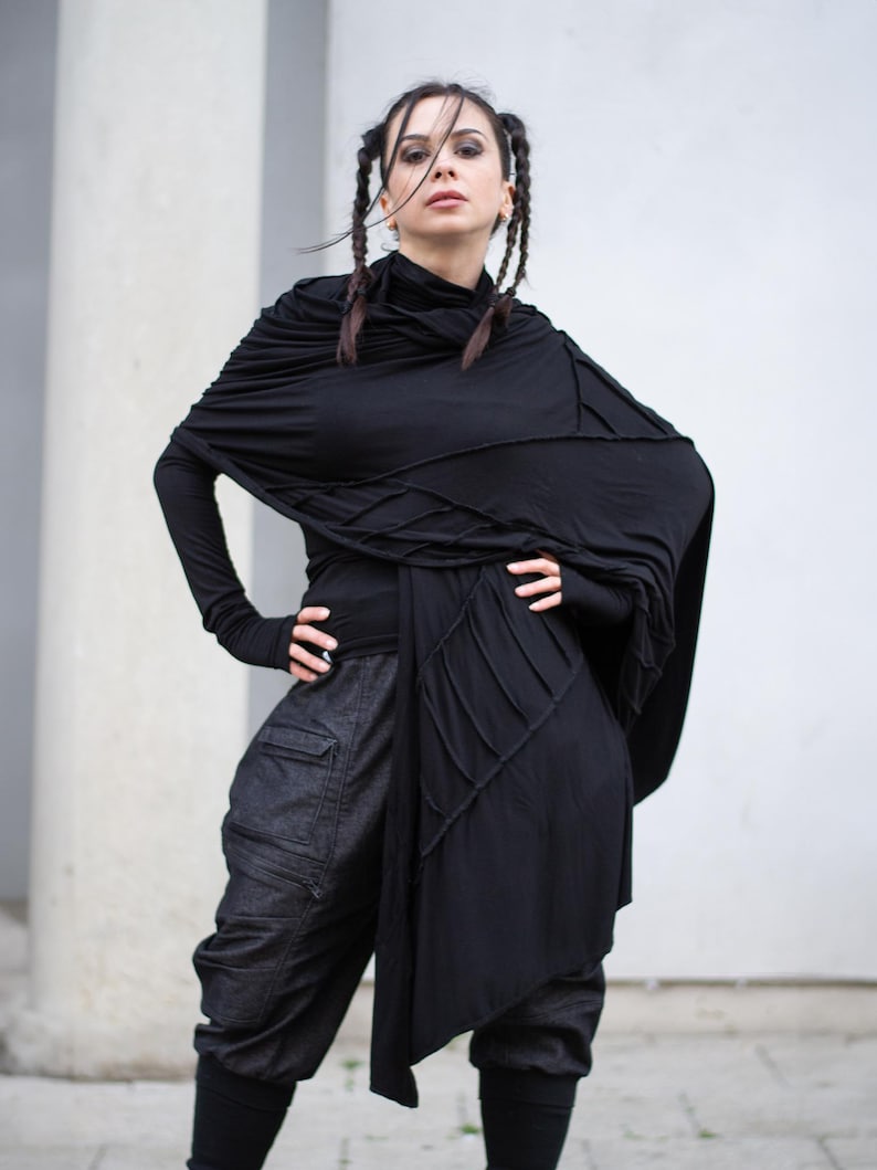 K&ouml;nnte beinhalten: Eine Frau in einem schwarzen Ensemble. Das Outfit umfasst ein lang&auml;rmliges Oberteil, einen drapierten Schal und eine dunkle Cargo-Hose aus Denim. Der Schal hat ein einzigartiges Nahtdesign. Die Frau hat dunkles Haar, das in Z&ouml;pfen gestylt ist.