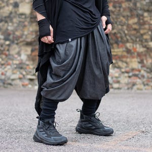 Może przedstawiać: Osoba nosząca czarny top, ciemnoszare spodnie haremki i czarne wysokie trampki. Spodnie mają unikalny, drapowany design. Strój jest ustawiony na tle ceglanej ściany i asfaltowej nawierzchni.