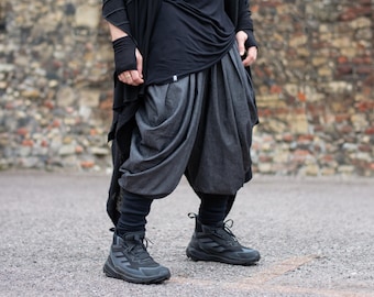 以下が含まれることがあります： 黒いトップス、濃いグレーのハーレムパンツ、黒のハイカットスニーカーを履いた人。 パンツはユニークなドレープデザインです。 レンガの壁とアスファルトの表面を背景にした服装です。