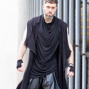 May include: A black sleeveless draped top and long vest are worn over a black top and black leather-look pants. The outfit includes black fingerless gloves. The background features a white wall and a metal gate.