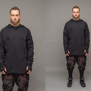 May include: A person wearing a black hooded sweatshirt with long sleeves and black trousers with red stitching. The person is standing in front of a grey background.