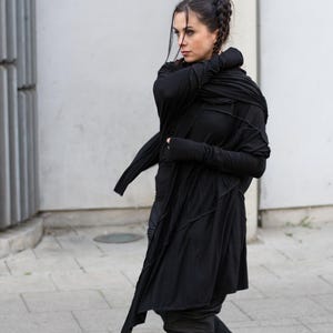 K&ouml;nnte beinhalten: Ein langer schwarzer Mantel mit asymmetrischen Eins&auml;tzen und einem drapierten Ausschnitt. Der Mantel wird mit schwarzen Leggings und Combat-Stiefeln kombiniert. Das Outfit hat eine dunkle, gotische &Auml;sthetik. Die Person geht auf einer gepflasterten Oberfl&auml;che.