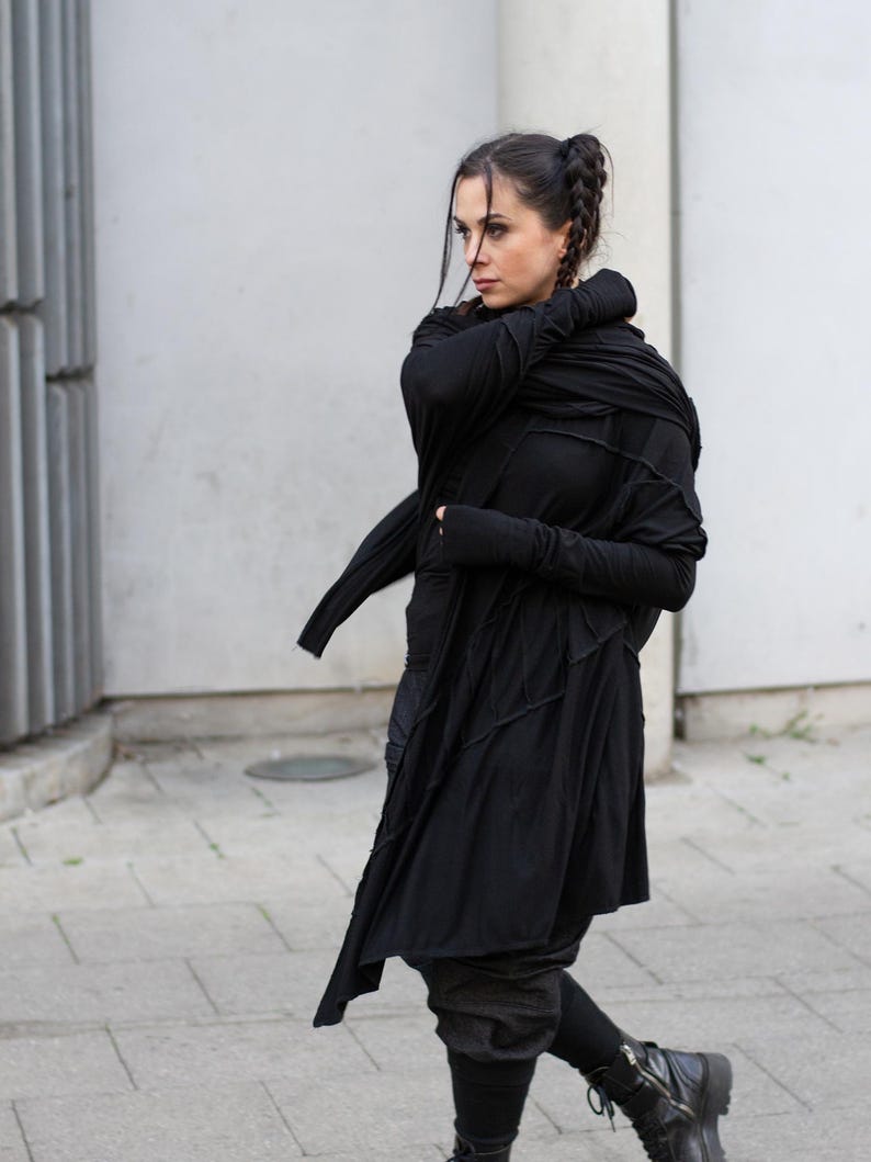 K&ouml;nnte beinhalten: Ein langer schwarzer Mantel mit asymmetrischen Eins&auml;tzen und einem drapierten Ausschnitt. Der Mantel wird mit schwarzen Leggings und Combat-Stiefeln kombiniert. Das Outfit hat eine dunkle, gotische &Auml;sthetik. Die Person geht auf einer gepflasterten Oberfl&auml;che.