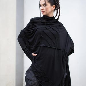 K&ouml;nnte beinhalten: Schwarzes Langarm-Oberteil mit hohem Ausschnitt und einzigartigem Nahtdesign. Das Oberteil hat eine drapierte, asymmetrische Silhouette. Das Model tr&auml;gt auch eine dunkle Jeanshose. Das Outfit ist ein moderner, kantiger Stil.