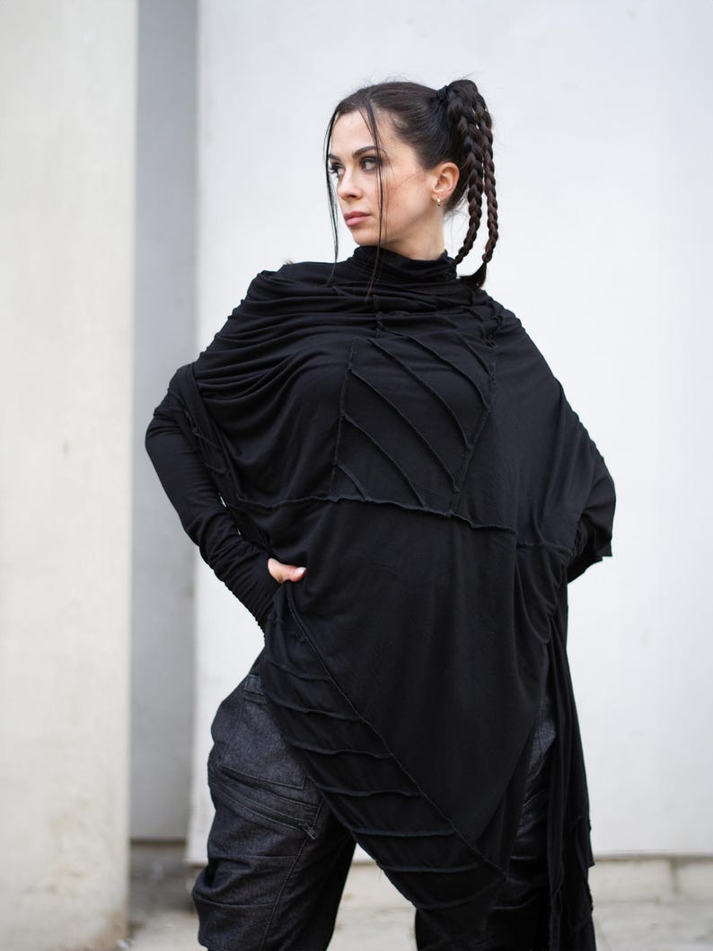 K&ouml;nnte beinhalten: Schwarzes Langarm-Oberteil mit hohem Ausschnitt und einzigartigem Nahtdesign. Das Oberteil hat eine drapierte, asymmetrische Silhouette. Das Model tr&auml;gt auch eine dunkle Jeanshose. Das Outfit ist ein moderner, kantiger Stil.