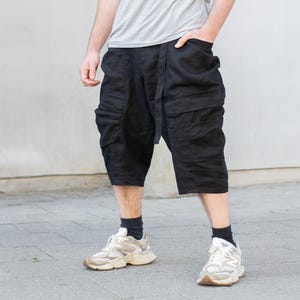 May include: Black shorts with multiple pockets and a belt. The shorts are knee-length and have a unique, layered design. The person is wearing white and beige sneakers and black socks. The background is a light gray wall and a gray sidewalk.