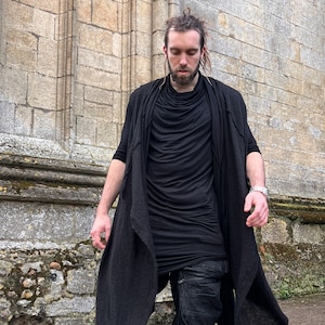 May include: A person wearing a black long-sleeved shirt, black pants, and a black long cardigan. The person is standing in front of a stone wall.