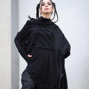 K&ouml;nnte beinhalten: Schwarzes Langarm-Top mit hohem Ausschnitt und asymmetrischem Design. Das Oberteil hat aufgen&auml;hte Details und eine drapierte, flie&szlig;ende Silhouette. Das Model tr&auml;gt eine dunkle Jeanshose. Das Outfit hat eine gotische oder alternative &Auml;sthetik.