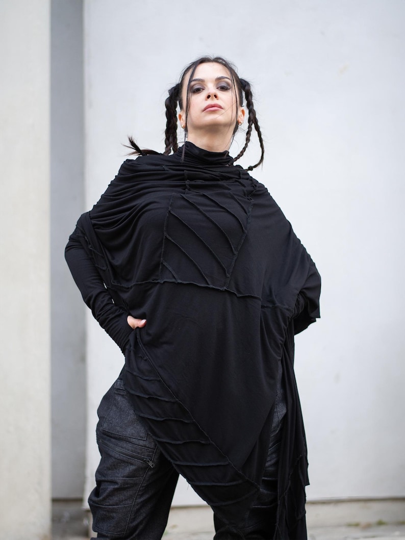 K&ouml;nnte beinhalten: Schwarzes Langarm-Top mit hohem Ausschnitt und asymmetrischem Design. Das Oberteil hat aufgen&auml;hte Details und eine drapierte, flie&szlig;ende Silhouette. Das Model tr&auml;gt eine dunkle Jeanshose. Das Outfit hat eine gotische oder alternative &Auml;sthetik.