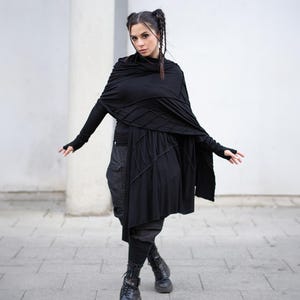 K&ouml;nnte beinhalten: Schwarzes Ensemble mit einem drapierten Schal, einem lang&auml;rmligen Oberteil und einem asymmetrischen Rock. Das Outfit umfasst Cargohosen und Kampfstiefel. Die Kleidung hat strukturierte Details und ein geschichtetes Design.