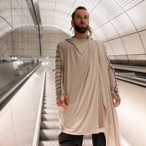 Puede incluir: Una persona que lleva una larga prenda con capucha de color crema con detalles negros en las mangas, de pie en una escalera mecánica en una moderna estación de metro con azulejos blancos.