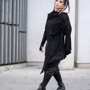 K&ouml;nnte beinhalten: Ganzk&ouml;rperaufnahme einer Person in einem schwarzen Outfit. Das Ensemble umfasst ein lang&auml;rmliges Oberteil mit asymmetrischem Design, dunkle Hosen, Leggings und schwarze Combat-Stiefel. Die Person geht auf einer gepflasterten Oberfl&auml;che.