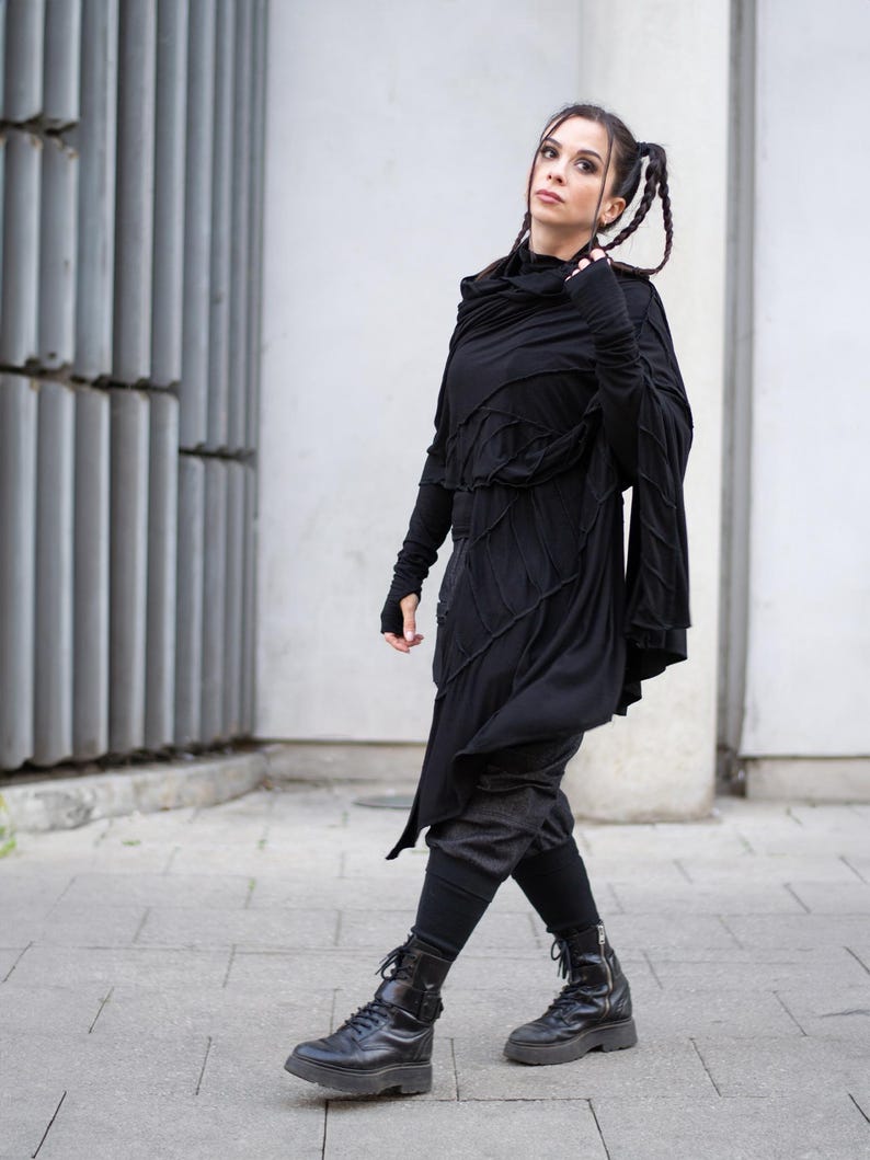 K&ouml;nnte beinhalten: Ganzk&ouml;rperaufnahme einer Person in einem schwarzen Outfit. Das Ensemble umfasst ein lang&auml;rmliges Oberteil mit asymmetrischem Design, dunkle Hosen, Leggings und schwarze Combat-Stiefel. Die Person geht auf einer gepflasterten Oberfl&auml;che.